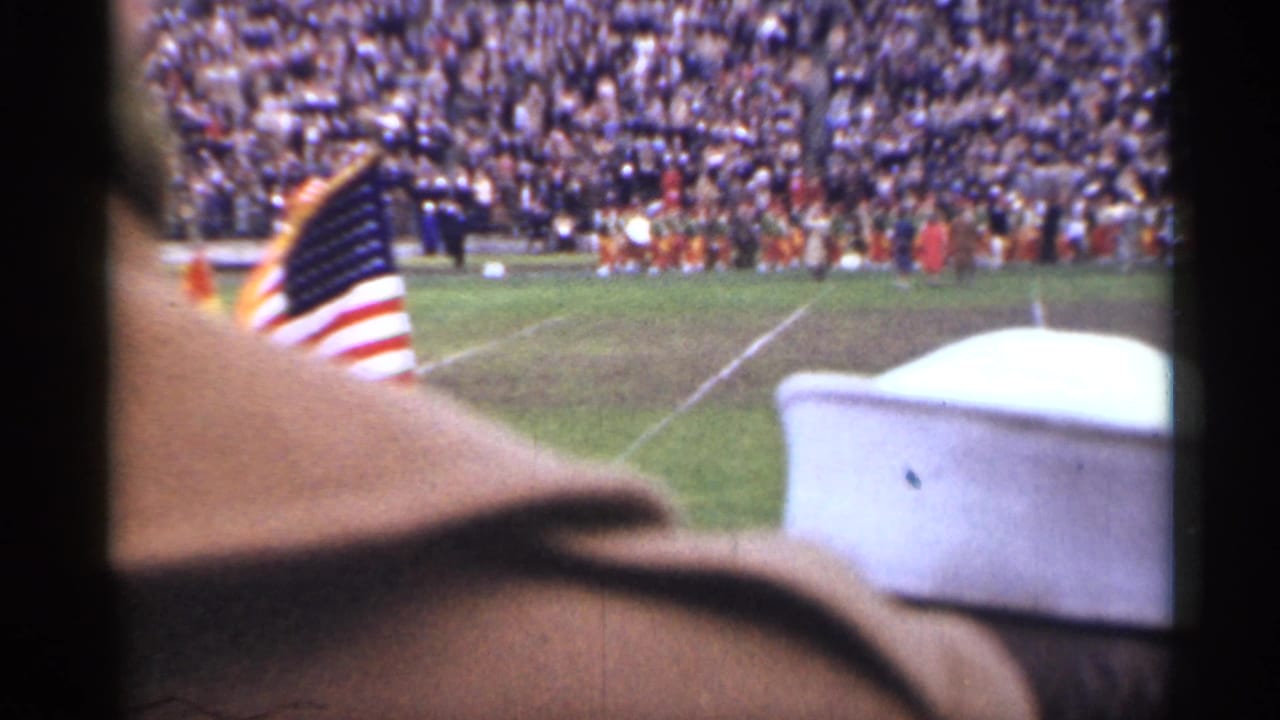 American flag and sailor cap in foreground with a marching band on a crowded stadium field.