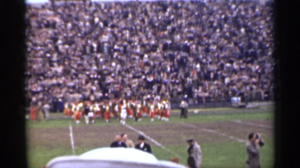 Home-movie frame from a 1944 San Francisco stadium event, with the band at mid-field and men in period hats near the camera.