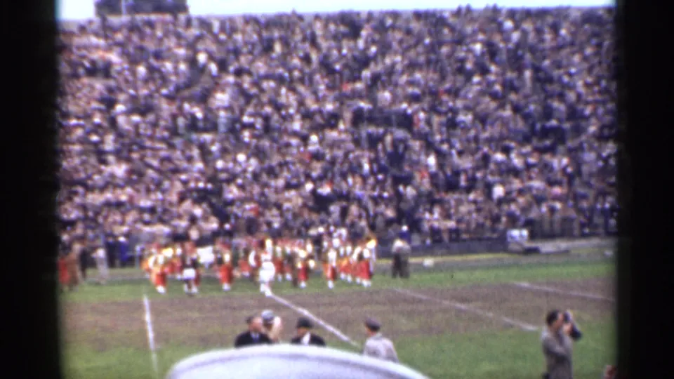 1944 San Francisco home-movie view of a marching band on the field, with spectators, a photographer, and a white cap in the foreground.