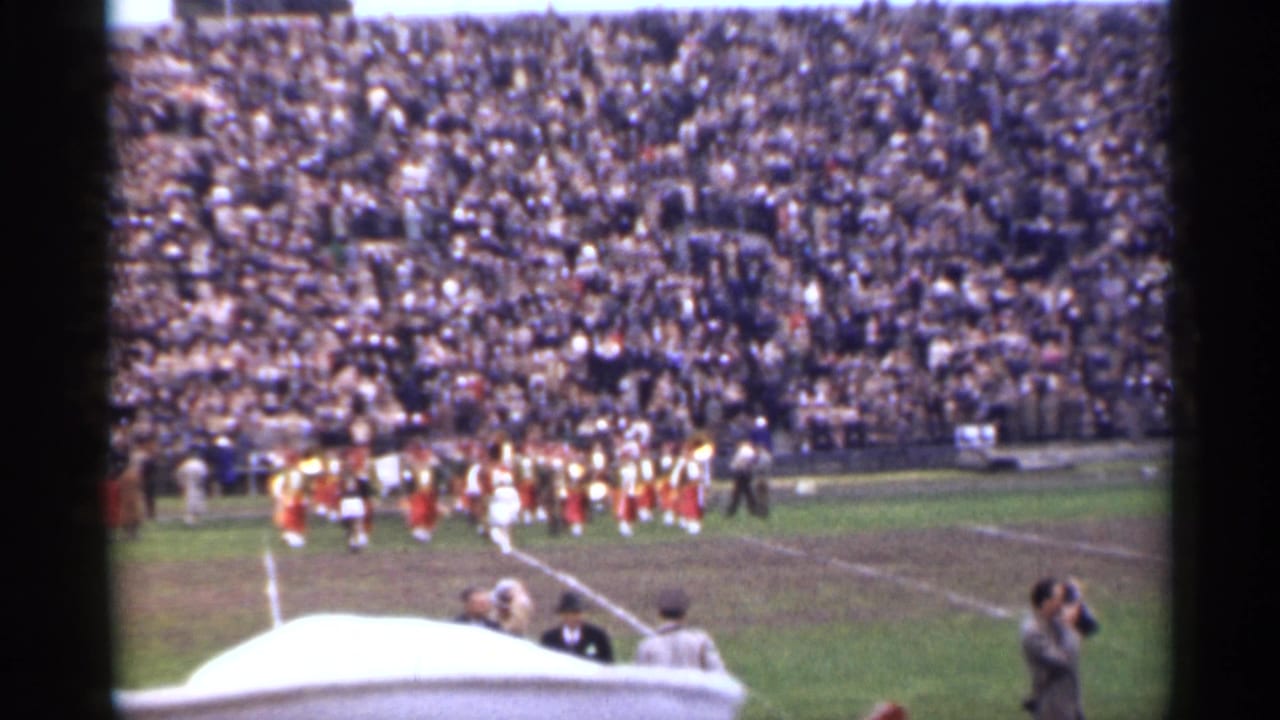 Packed stadium with a marching band on the field and spectators near the sideline.