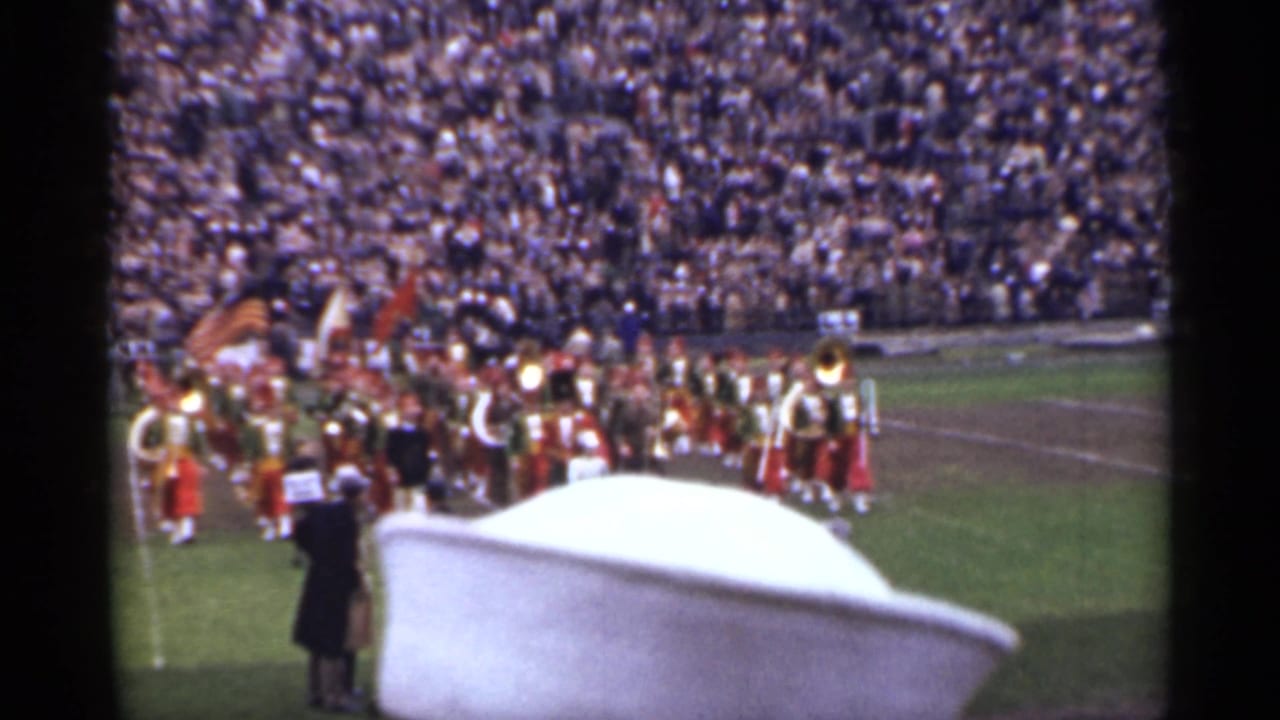 Marching band on a football field before a crowded stadium, partly blocked by a white hat.