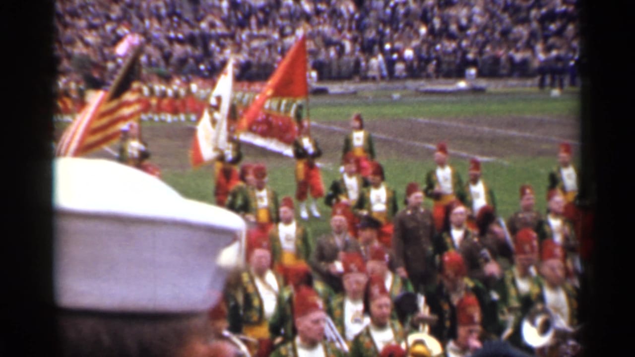 Marching band with flags on a football field before a crowded stadium.