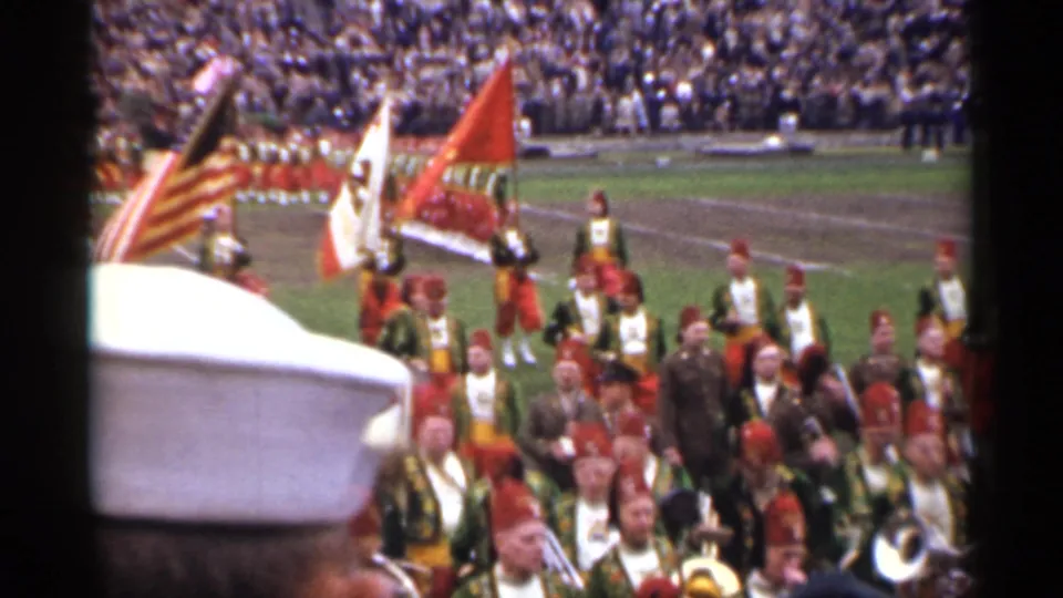 1944 San Francisco home-movie frame showing a marching band in red and green uniforms carrying flags on a football field.