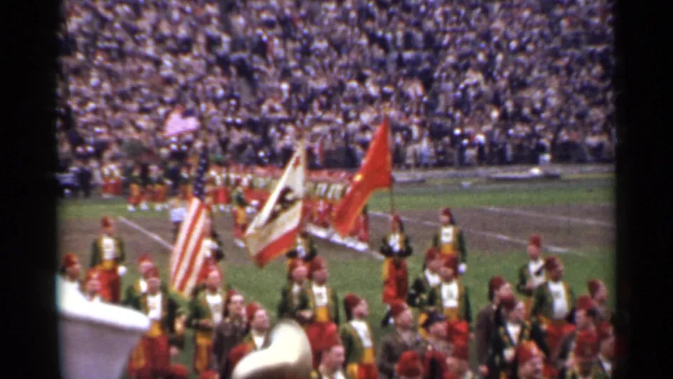1944 San Francisco 8mm frame with band members in ornate uniforms, flags raised, and a dense stadium crowd in the background.