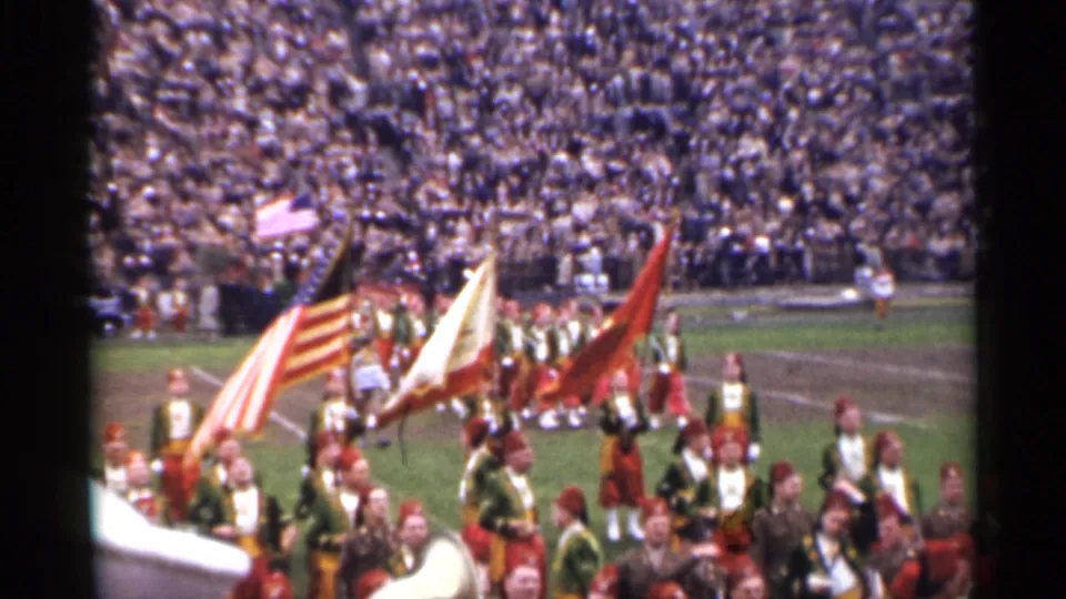 1944 San Francisco 8mm frame showing a marching band, raised flags, and a packed stadium during a field ceremony.