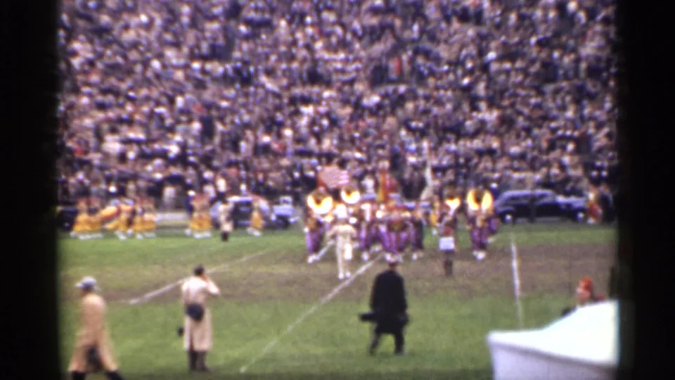 1944 San Francisco home-movie frame showing marching musicians on a stadium field with spectators and sideline cars in view.