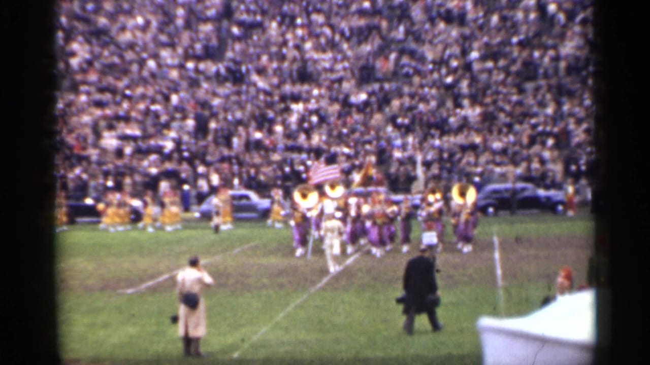 Marching band enters a football field as spectators fill the stadium stands.