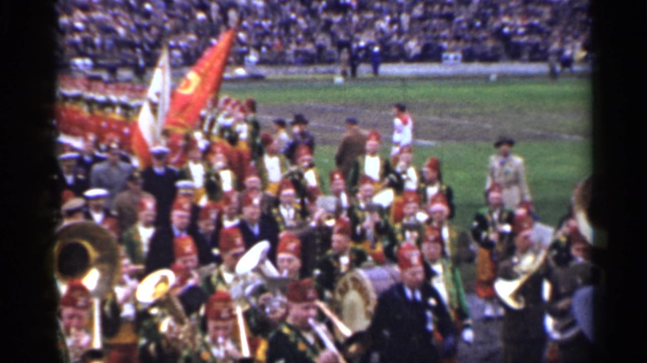 Marching band and flag bearers pass in front of crowded stadium stands.