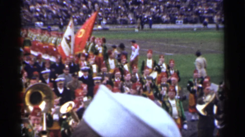 Uniformed band and flag bearers continue past the camera at a San Francisco stadium, with spectators and field activity behind them.