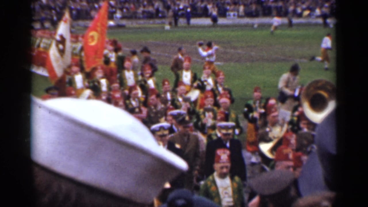 1944 San Francisco home-movie frame showing a marching band on a football field with spectators and foreground hats partially blocking the view.
