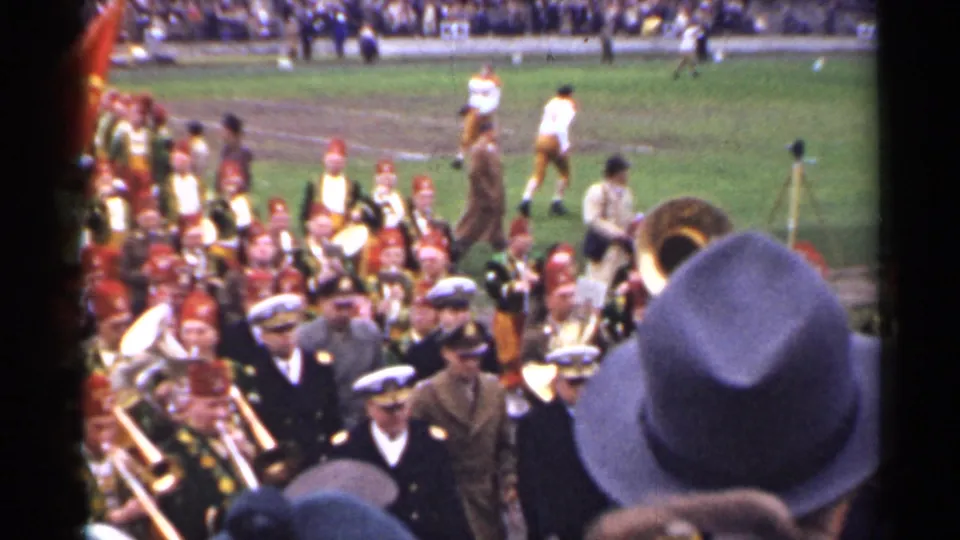 1944 San Francisco frame with a marching band and uniformed figures near the sideline while football players stand in the background.