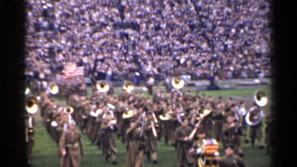 Wide 1944 San Francisco frame showing a marching band crossing the football field before crowded stadium stands and an American flag.