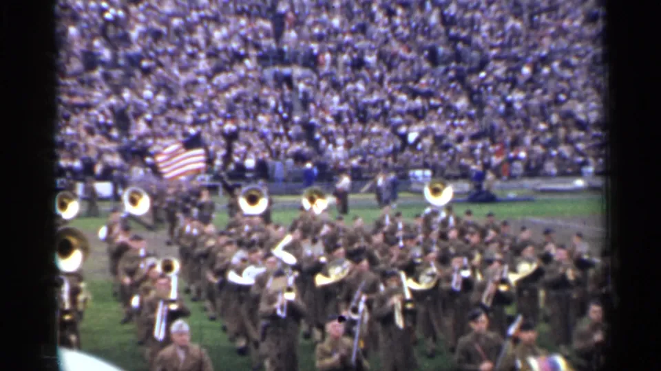 Military musicians in brown uniforms carry brass instruments and an American flag during the stated 1944 San Francisco stadium performance.