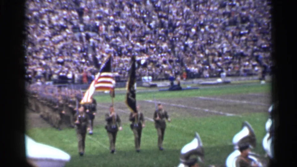 Flag bearers, including an American flag, march on a San Francisco football field during a 1944 military band sequence.