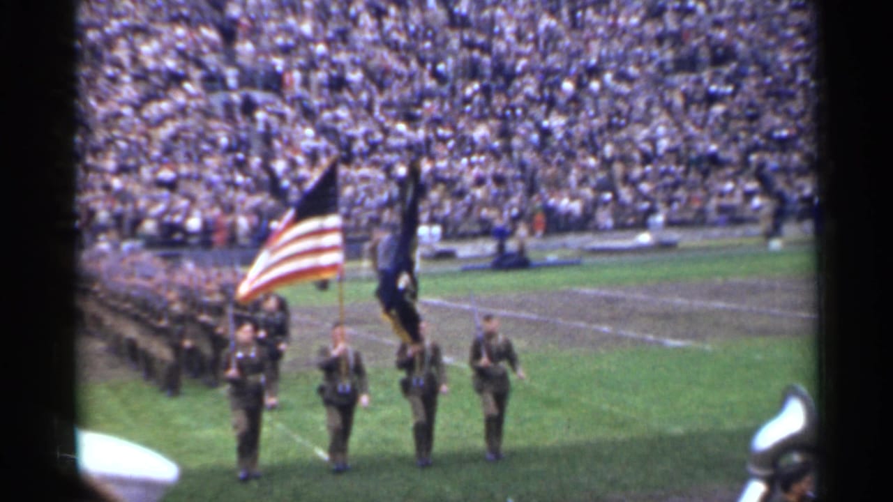 Color guard with American flag and dark standard on a stadium field before a large crowd.