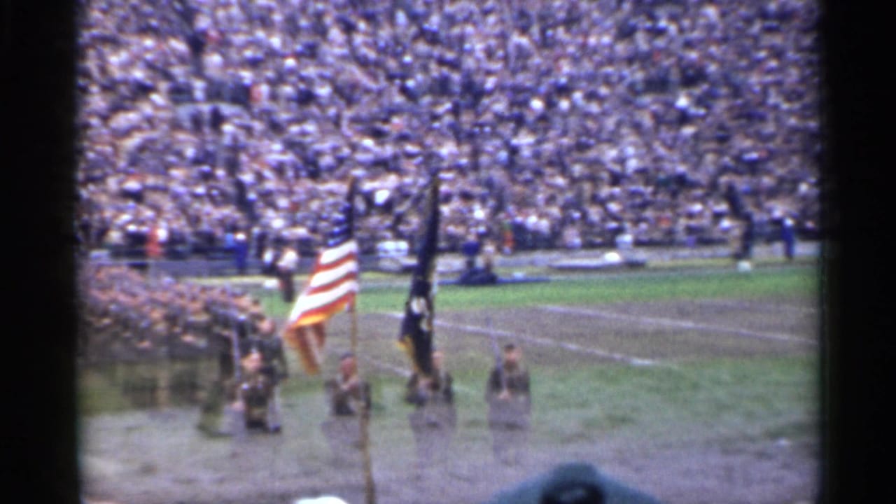 Military flag bearers appear on a San Francisco stadium field in 1944, filmed from the spectator area with crowded stands behind.