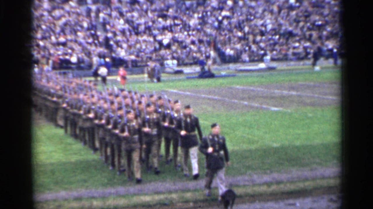 Soldiers march in formation across a stadium field as spectators fill the stands.