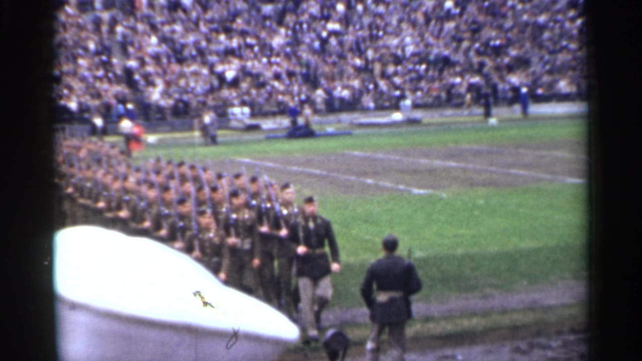 Uniformed troops continue marching through the stadium field in 1944 San Francisco footage, partly obscured by a pale foreground object.