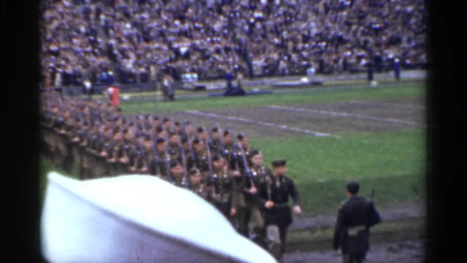 The spectator-shot 8mm frame shows soldiers in formation at a San Francisco stadium, with a foreground obstruction and a packed crowd beyond.