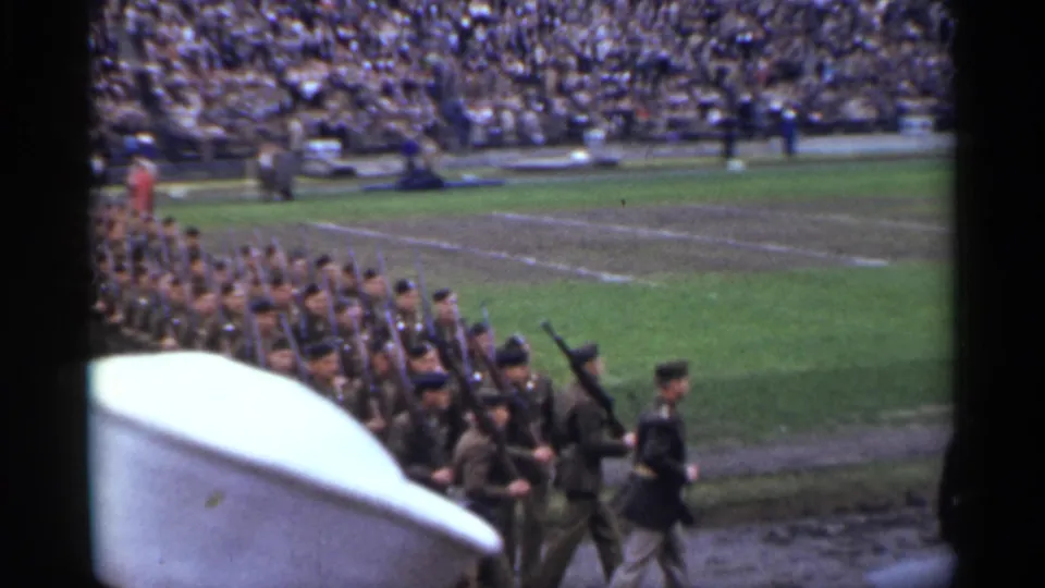 A military parade formation moves through the San Francisco stadium frame, with rifle-bearing soldiers, an officer, and spectators in the stands.