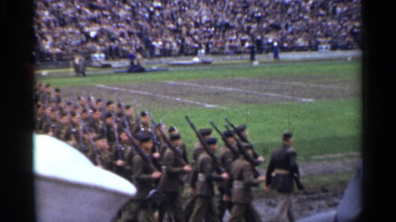 Soldiers with rifles march past a crowded stadium from an elevated spectator view.