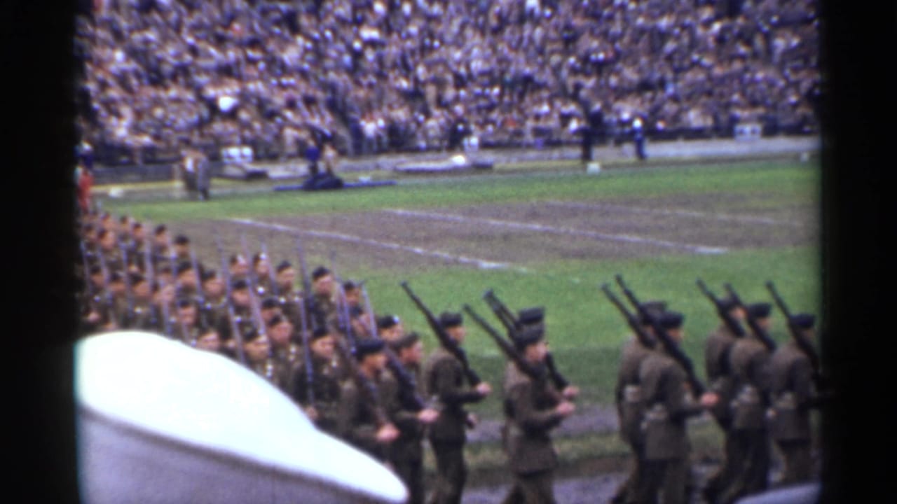 Uniformed troops with shouldered rifles pass through a crowded San Francisco stadium during the 1944 parade footage.