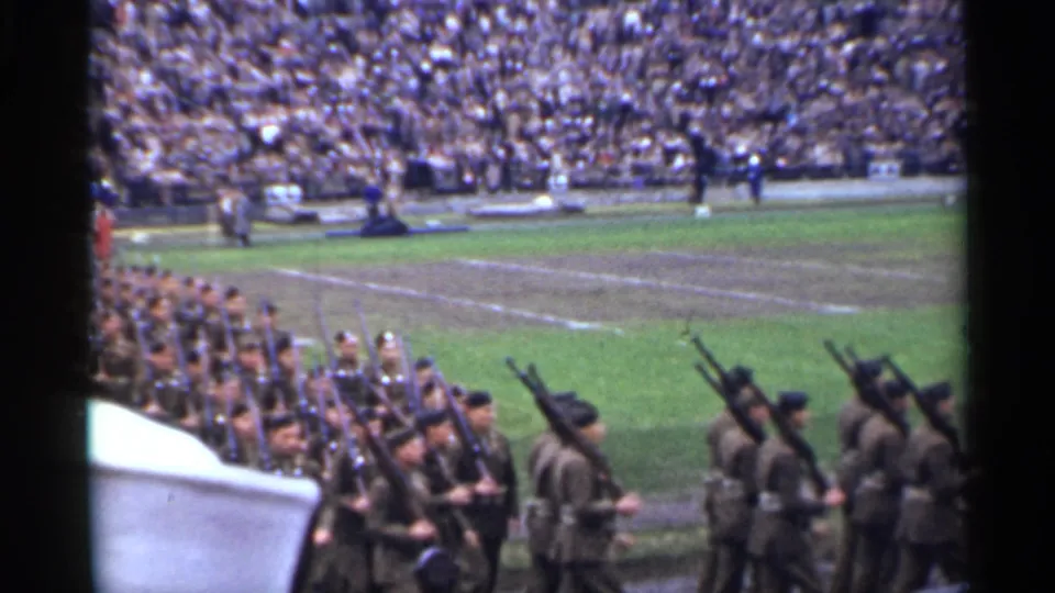 A line of soldiers with rifles advances through the stadium parade route, filmed from a high spectator position.