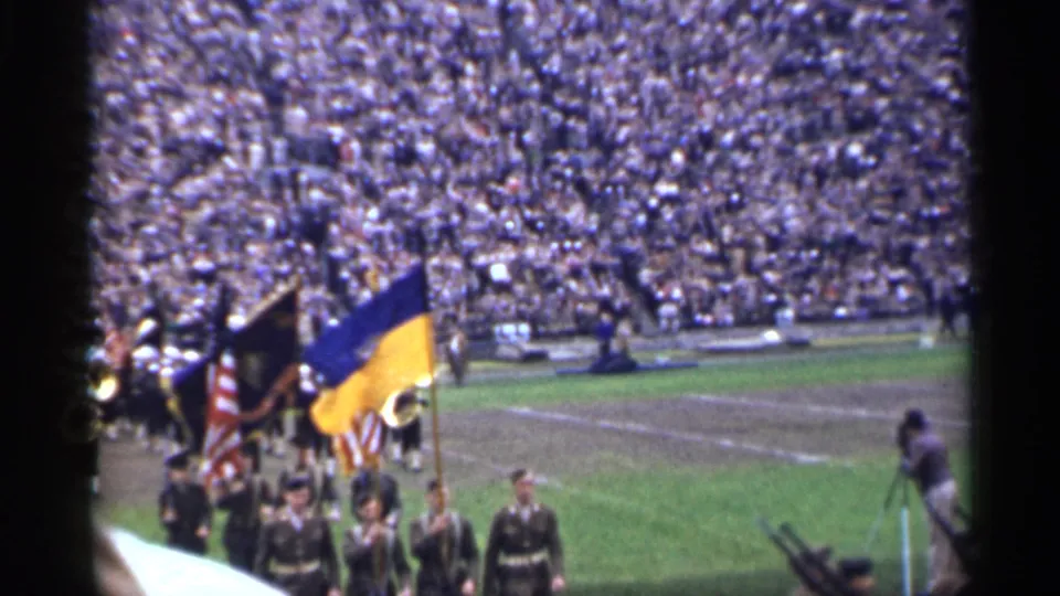 Flag bearers, including a blue and yellow flag, move onto the stadium field as soldiers continue marching nearby.