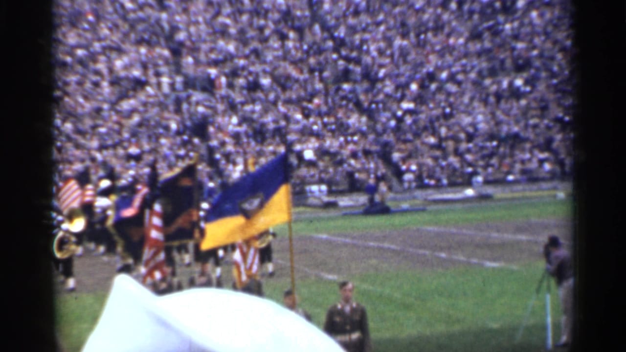 Color guard and marching band crossing a stadium field before crowded stands.