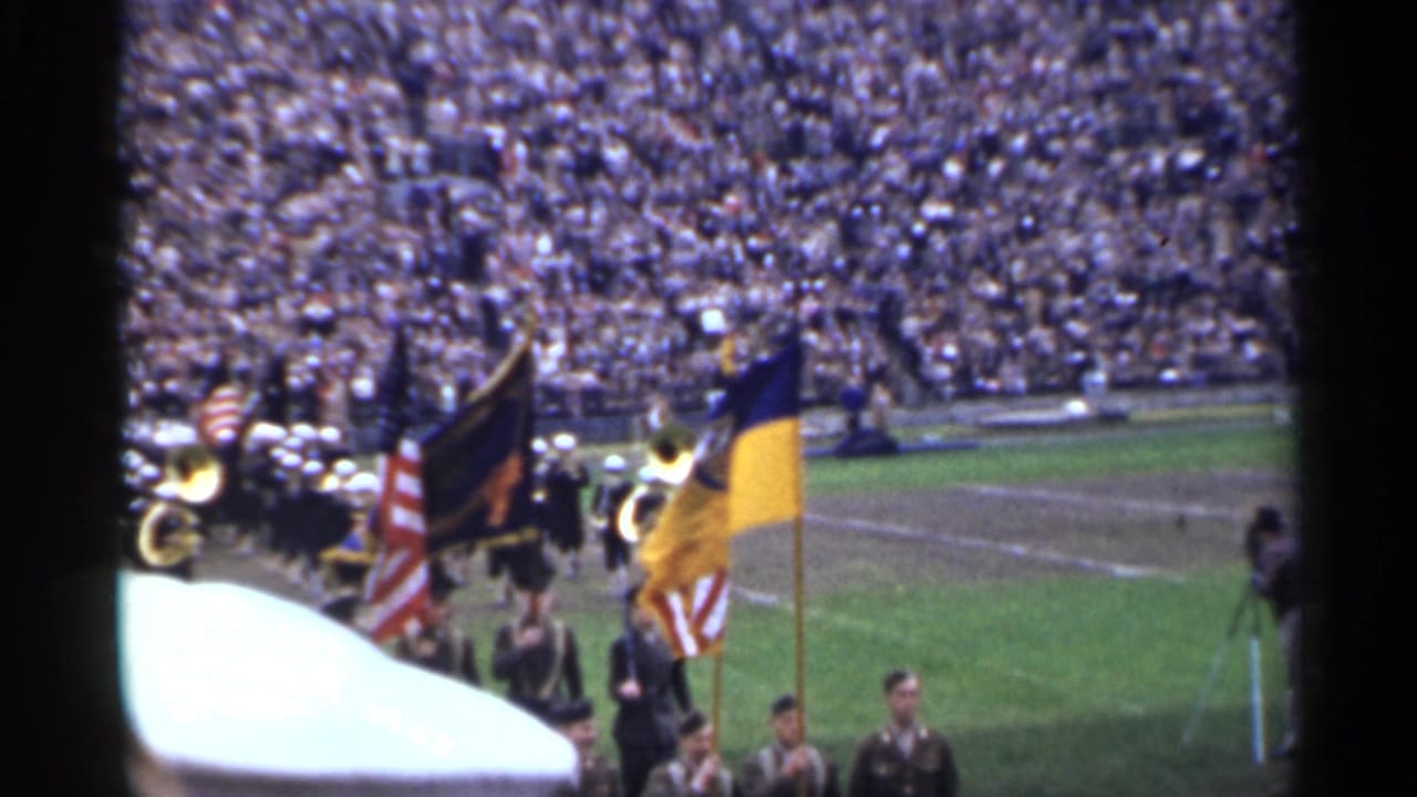 Uniformed color guard and brass band pass across a San Francisco stadium field in 1944 with spectators packed in the background.