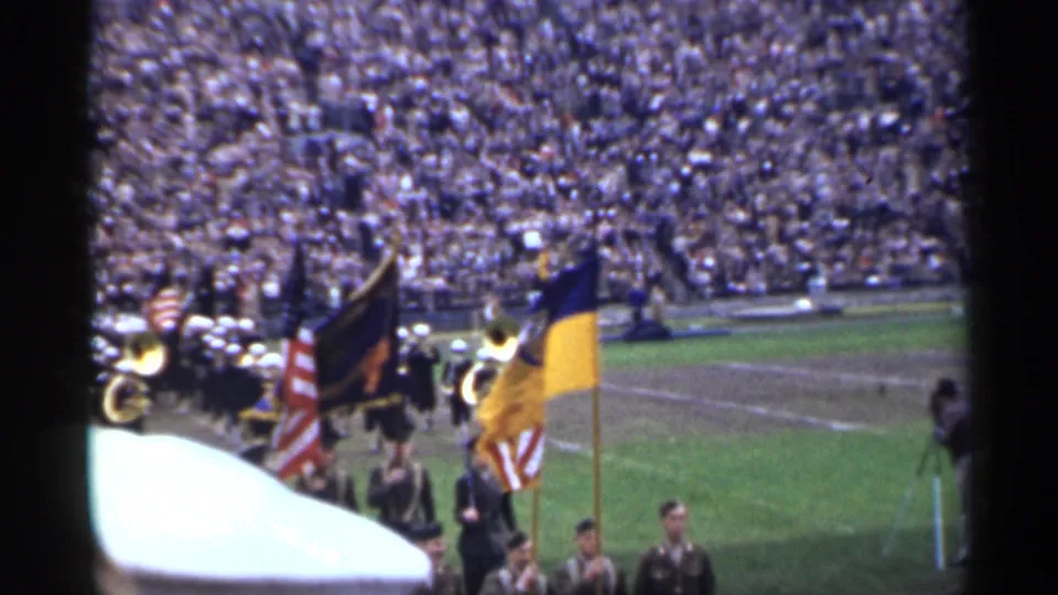 Uniformed color guard and brass band pass across a San Francisco stadium field in 1944 with spectators packed in the background.
