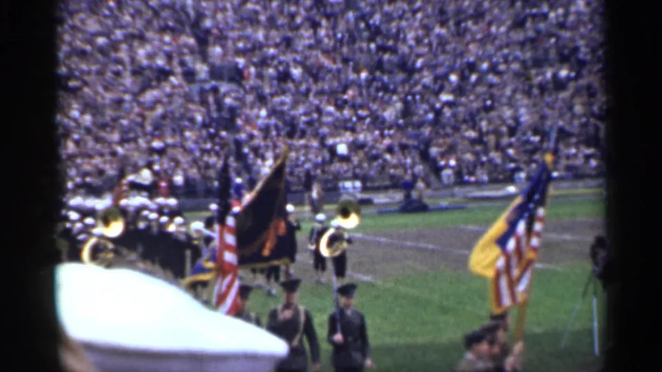 1944 home-movie view of soldiers or uniformed marchers carrying flags while brass musicians stand nearby on a crowded stadium field.