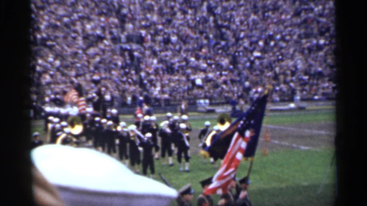 Marching band entering a football field with flags and a large crowd behind them.