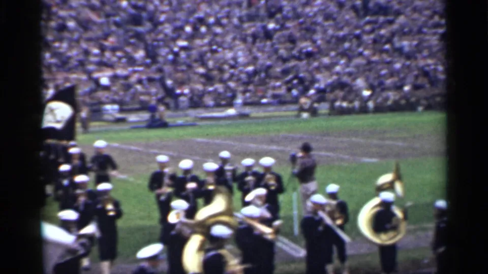 Uniformed band members with white caps and brass instruments cluster near the sideline in 1944 San Francisco stadium footage.