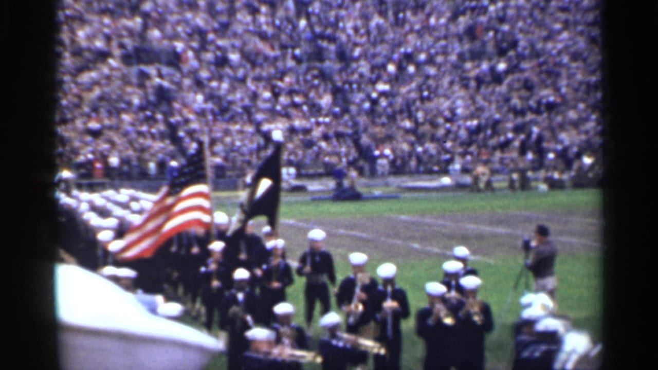Sailors march across a stadium field behind an American flag and dark standard.