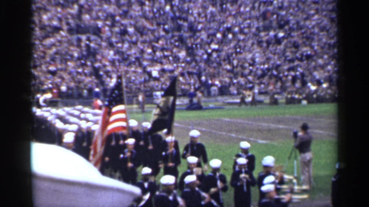 Sailors and band members pass through a stadium field in San Francisco, with an American flag, dark standard, and spectators filling the stands.