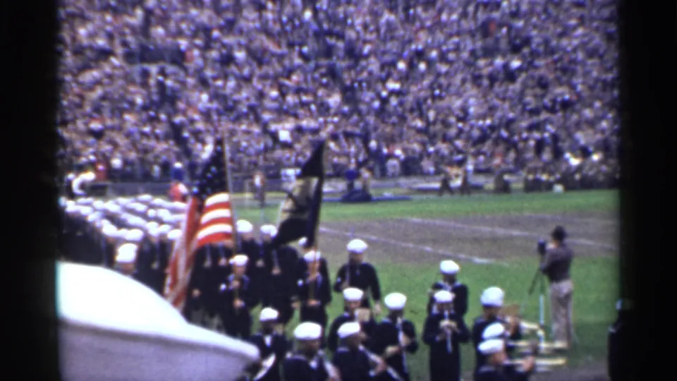 Sailors and band members pass through a stadium field in San Francisco, with an American flag, dark standard, and spectators filling the stands.