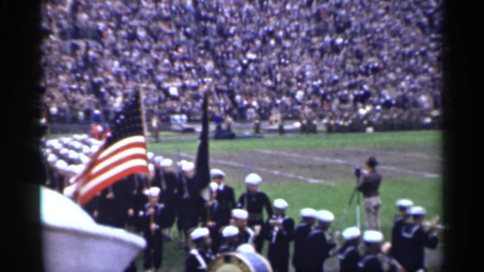 The American flag fills the foreground as sailors and musicians march through a crowded San Francisco stadium in 1944 archival home-movie footage.