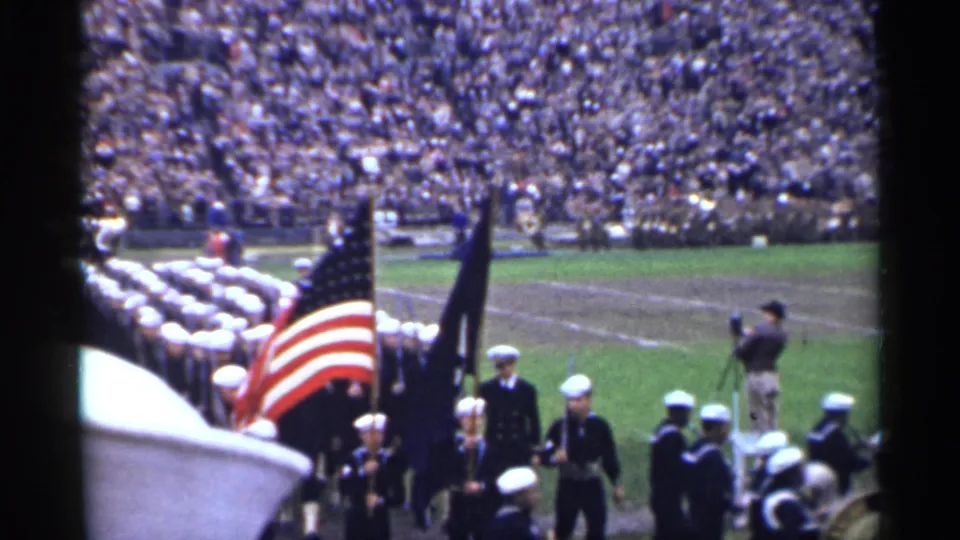Near the end of the clip, sailors, a U.S. flag, and a dark standard move past a field cameraman with packed stadium seating in the background.
