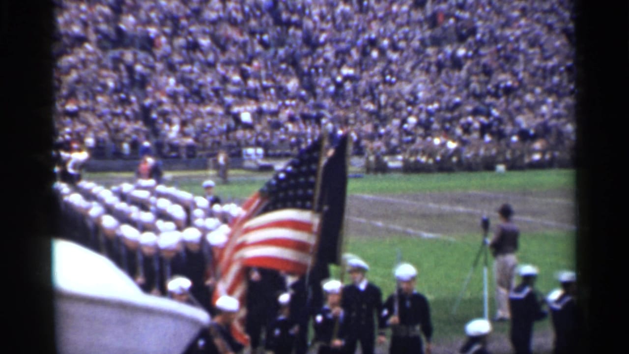 Sailors march on a field with an American flag before packed stadium stands.