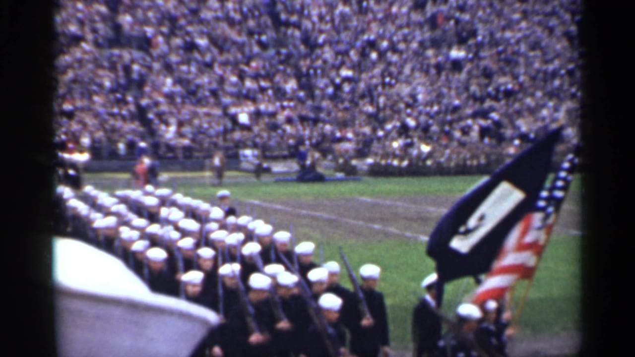Navy formation passes with rifles and flags on a stadium field.