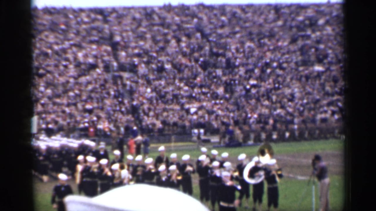 Marching band performs on a football field before packed stadium stands.