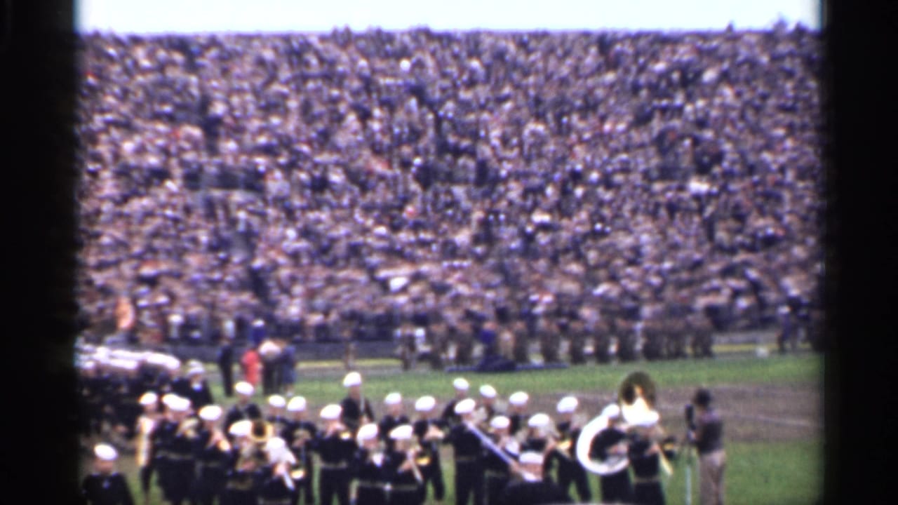 A marching band in dark uniforms and white hats performs on a San Francisco football field in 1944, with packed stands behind.