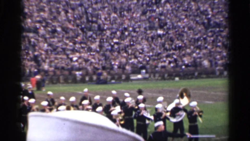 A dark-uniformed marching band with white hats moves across a 1944 San Francisco football field before a large stadium crowd.