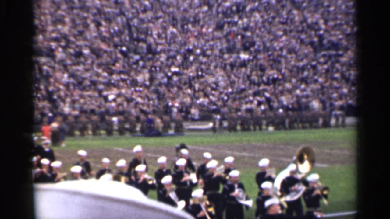 Military band members in white caps gather near a field as a packed stadium crowd fills the background.