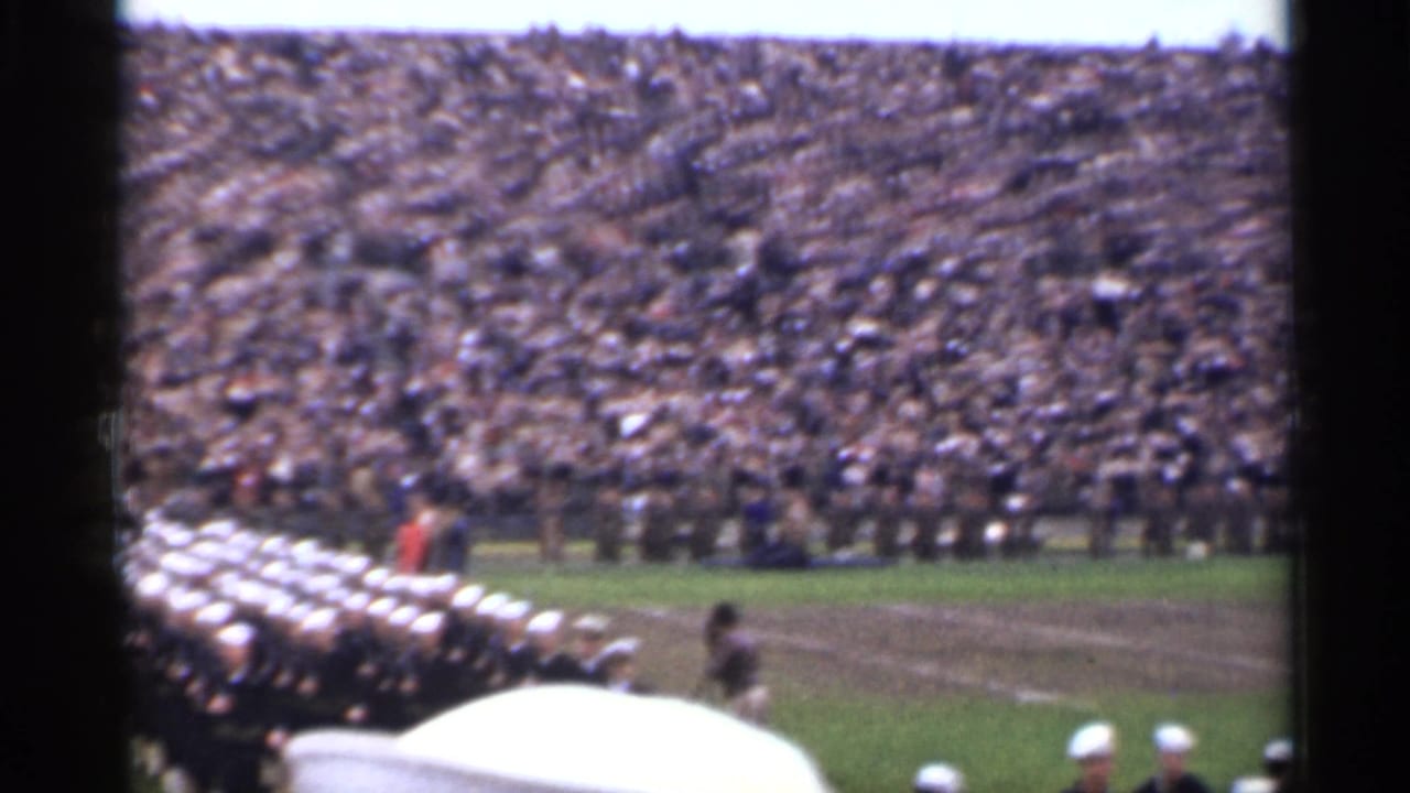 Uniformed formation crosses the field near crowded stadium seating.