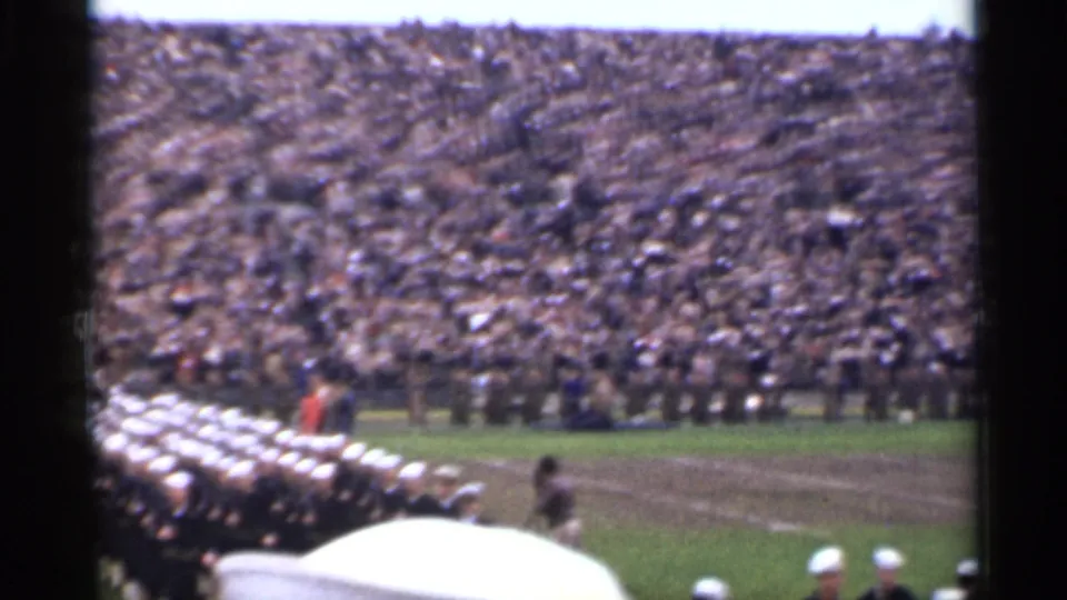 1944 San Francisco home-movie frame with a dark-uniformed marching formation and white caps in front of packed bleachers.