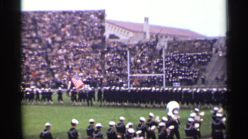 1944 San Francisco 8mm frame of band members in white caps performing on a football field beside an American flag and goalposts.