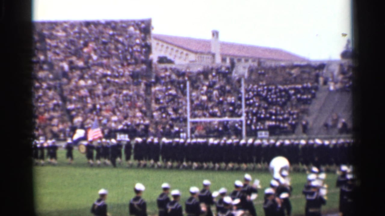 Marching band in formation on a football field with packed bleachers behind it.