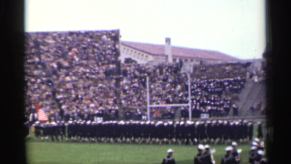 1944 San Francisco home-movie frame of a long marching-band line on a football field with spectators packed into the stands.